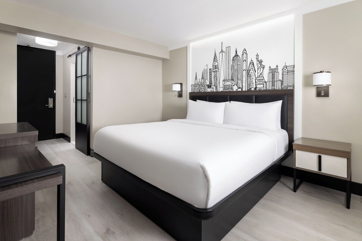 A modern hotel room with a large bed, a desk, a chair, and a cityscape wall art.