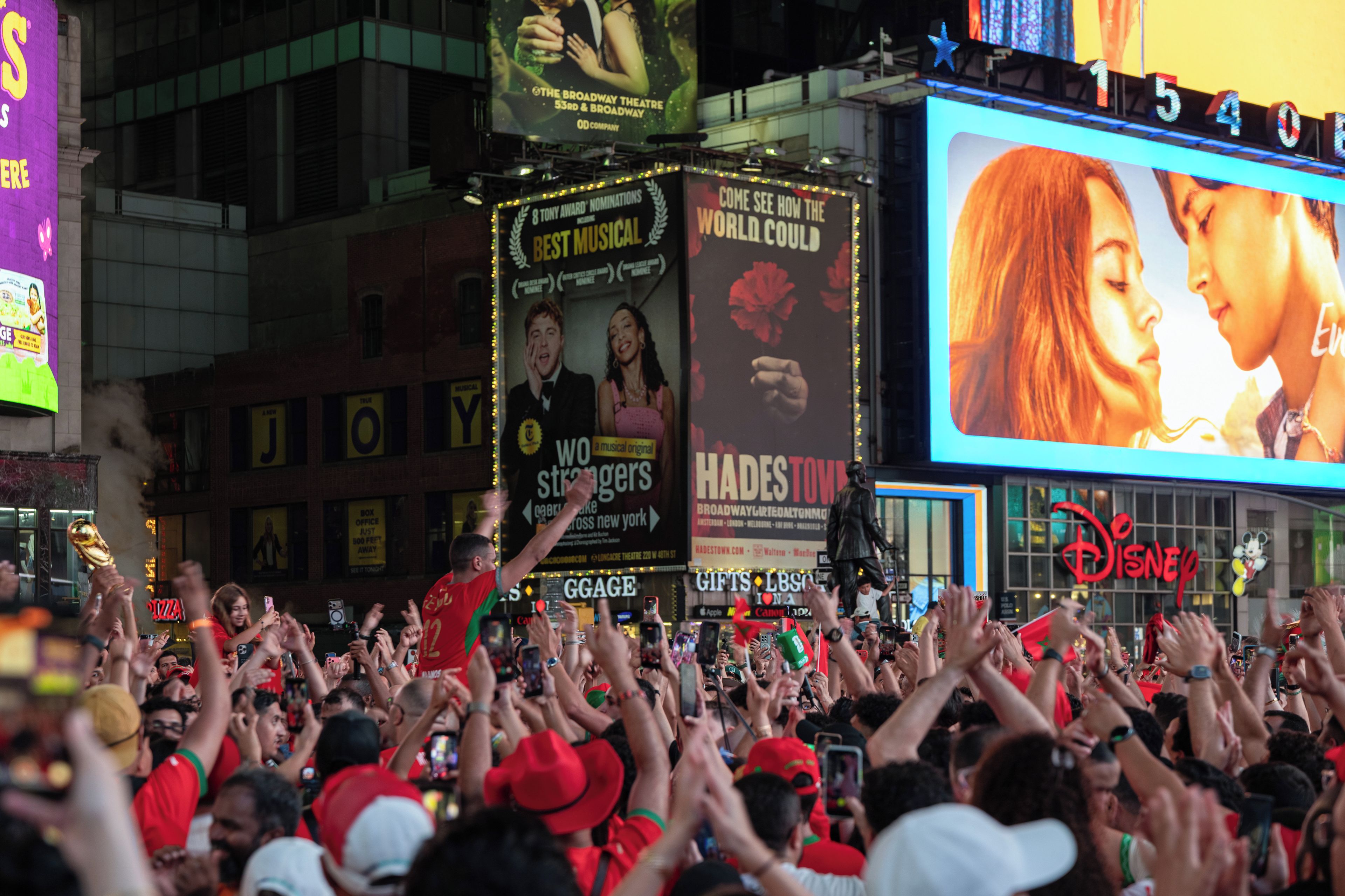 A crowded urban area with a large digital billboard advertising a musical.