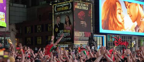 A crowded urban area with a large digital billboard advertising a musical.