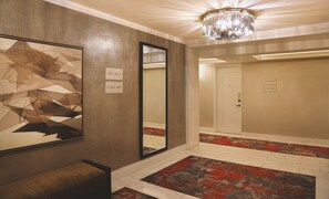 A hotel lobby with a geometric wall art, a mirror, a chandelier, a bench, and a red and gray patterned carpet.