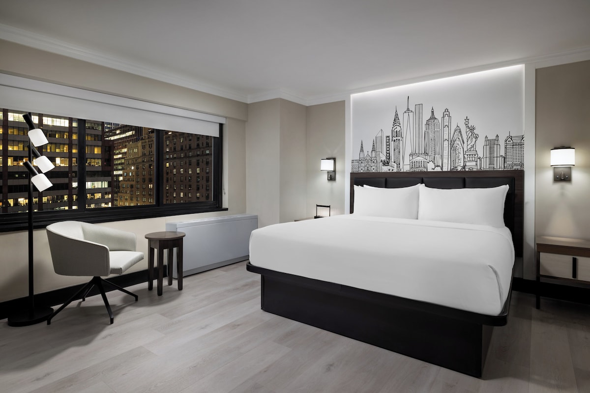 A modern hotel room with a large bed, a city skyline mural, a city view through the window, a small table, and a chair.
