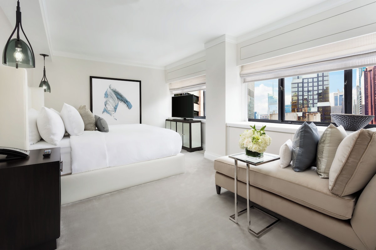 A modern hotel room with a large bed, a sofa, a TV, and a city view.