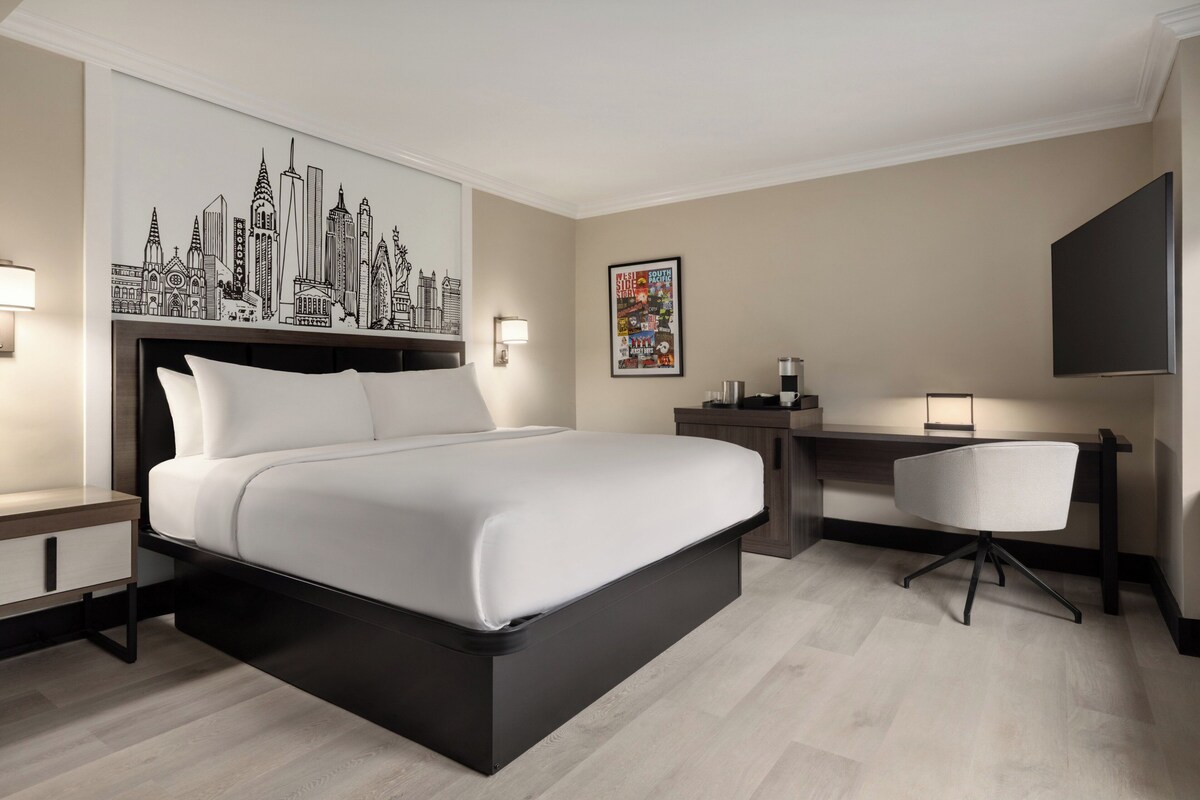 Habitación de hotel con una cama grande, un escritorio, una silla y una pared con vista a la ciudad y un cartel de cine.