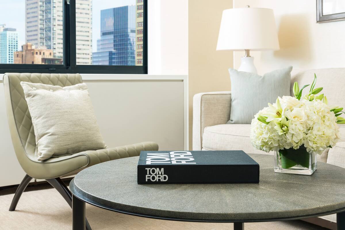 Een moderne zithoek met een ronde salontafel, een boek getiteld 'TOM FORD', een vaas met witte bloemen en een uitzicht op de stad door een raam.