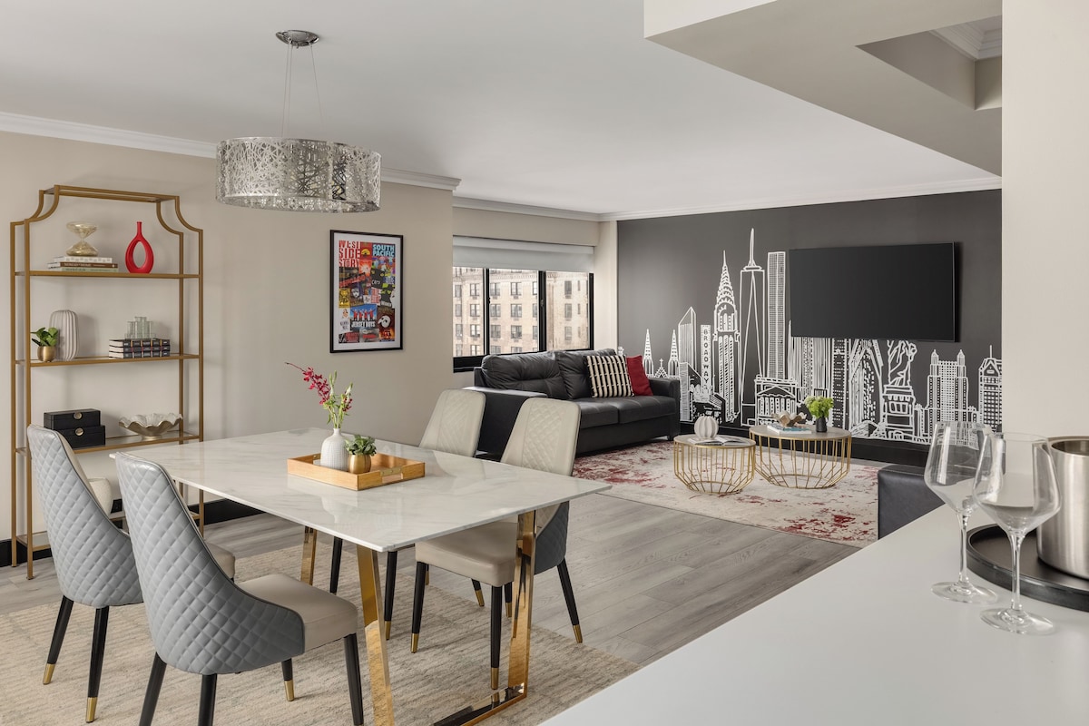 A modern living room with a dining area, a black sofa, a chandelier, and a cityscape mural on the wall.