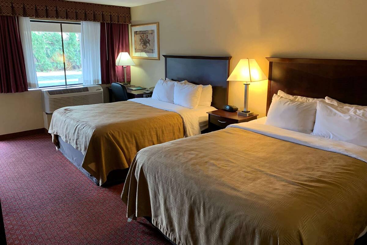 A hotel room with two beds, a window with curtains, an air conditioning unit, a desk with a lamp, and a framed picture on the wall.