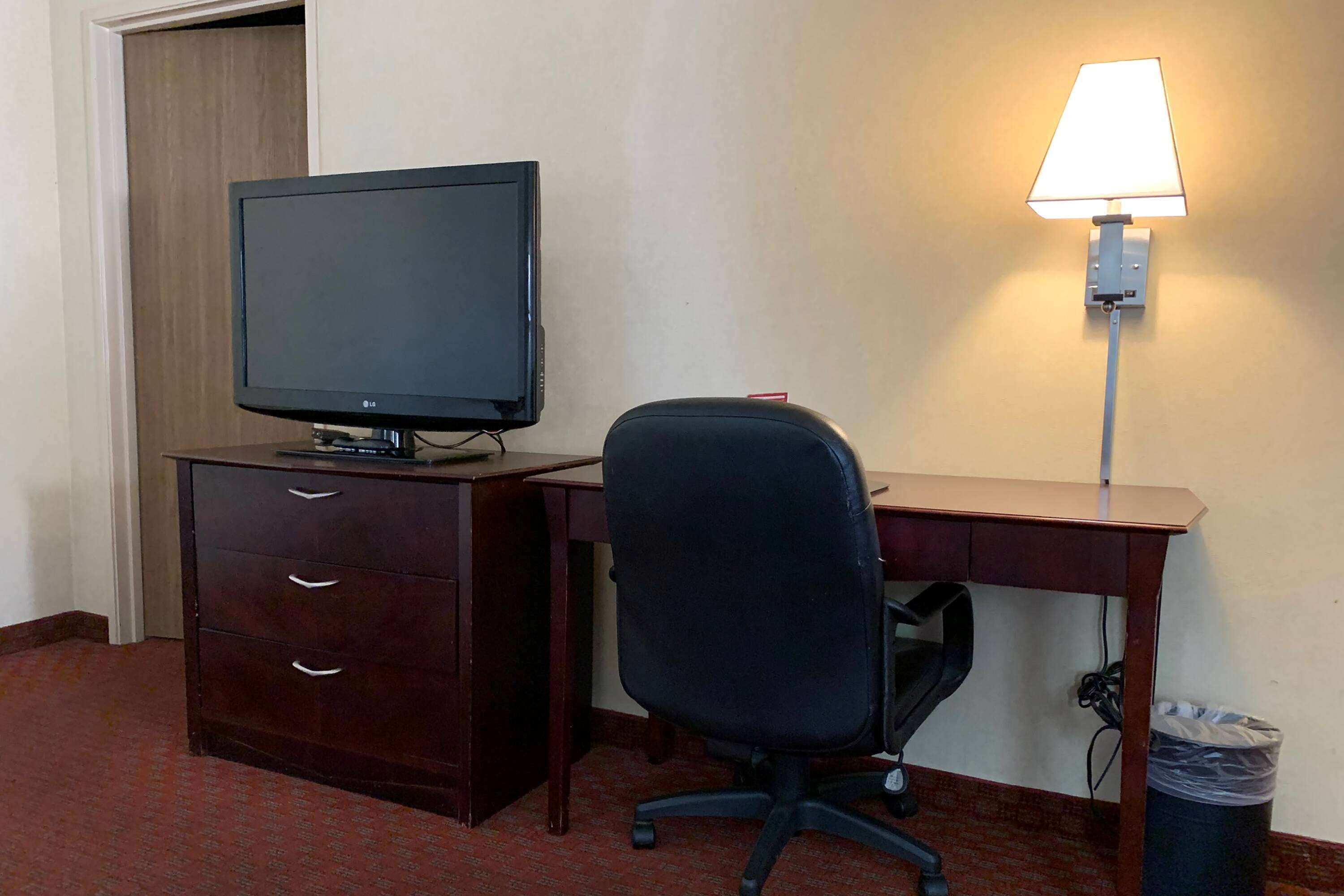 A hotel room with a desk, a chair, a television, and a wall-mounted lamp.