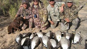 A group of people in camouflage attire posing with a dog and several birds on the ground.