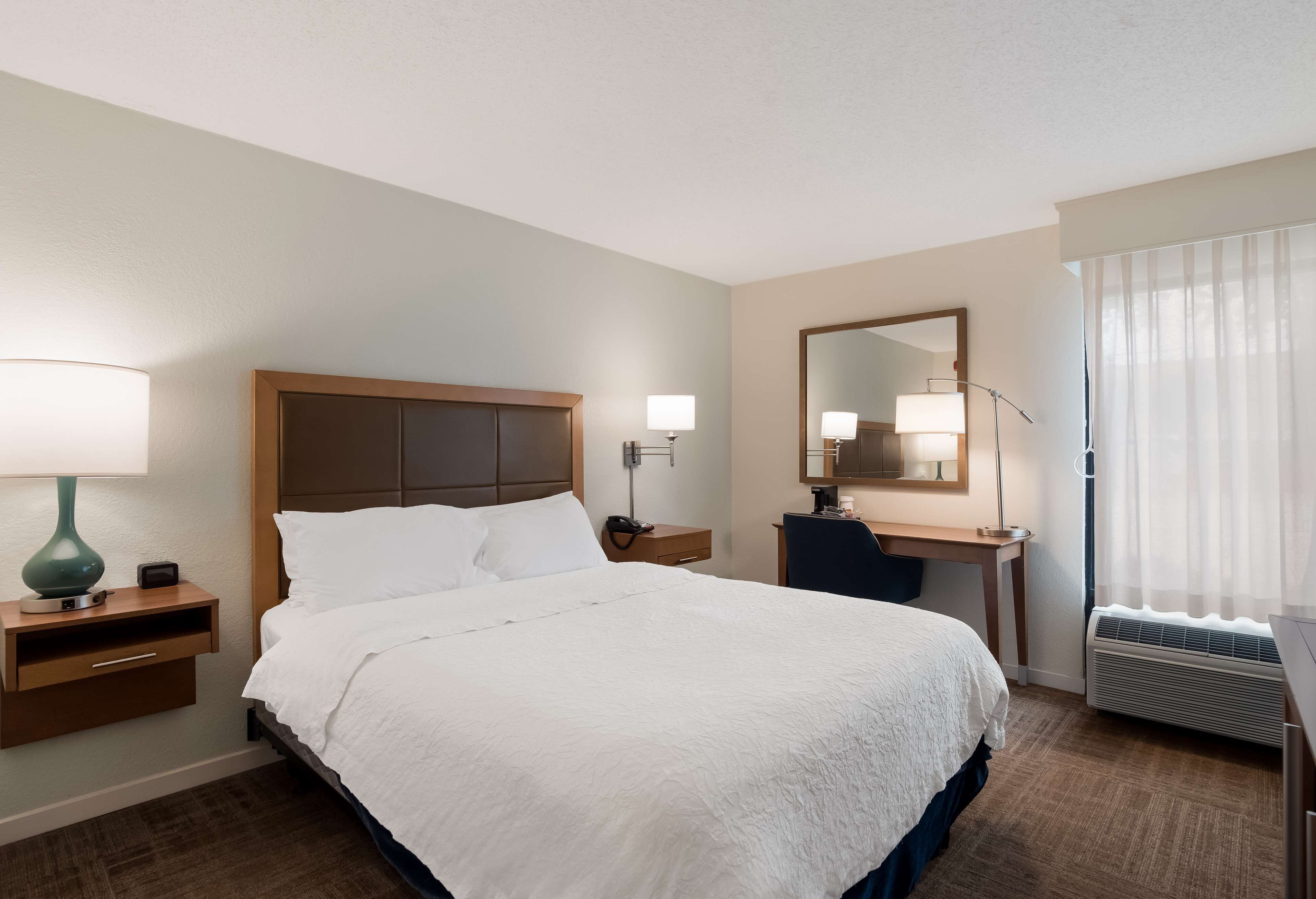 A hotel room with a large bed, bedside tables with lamps, a desk with a chair, a mirror, and a window with curtains.