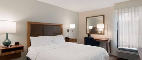 Quarto de hotel com uma cama grande, mesas de cabeceira com luminárias, uma escrivaninha com uma cadeira, um espelho e uma janela com cortinas.