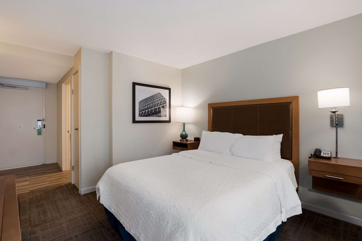 A hotel room with a large bed, a bedside table with a lamp, a framed picture on the wall, and a wooden nightstand.
