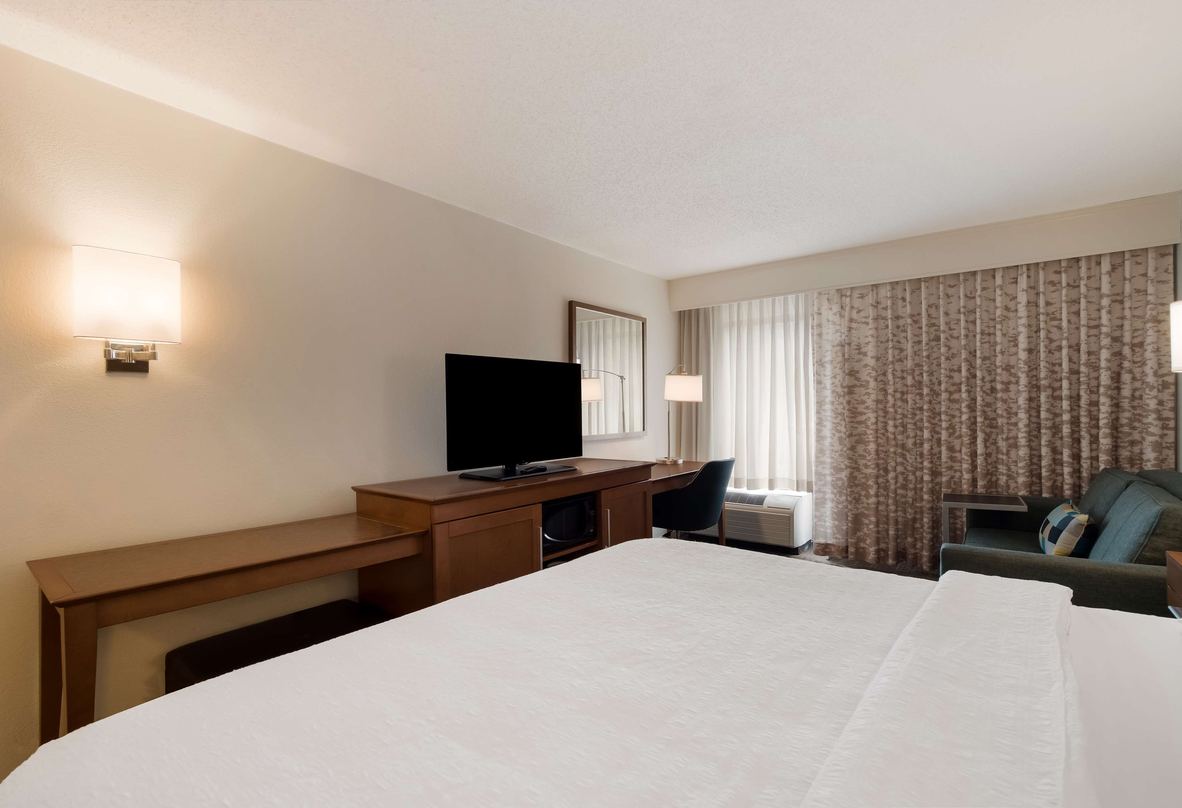 A hotel room with a large bed, a desk with a flat-screen TV, a chair, and a window with curtains.