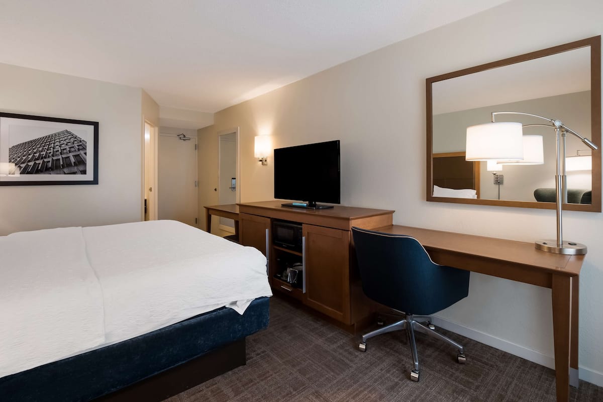 A hotel room with a bed, a desk with a computer, a chair, a TV, and a mirror.