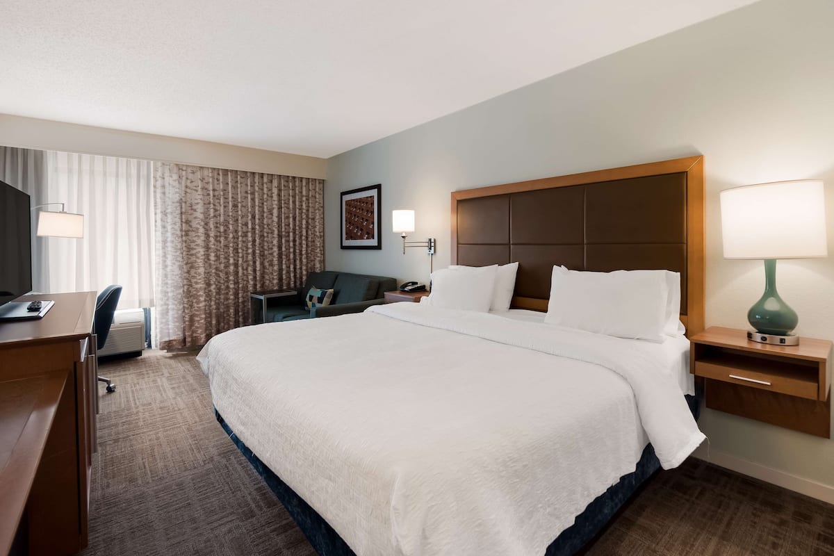 A hotel room with a large bed, a desk with a chair, a small table, and a lamp.
