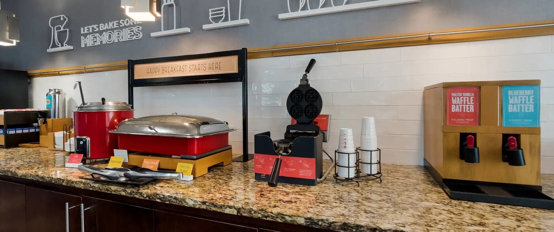 A breakfast bar with waffle batter dispensers, a waffle iron, and a sign that reads "HAPPY BREAKFAST STARTS HERE".