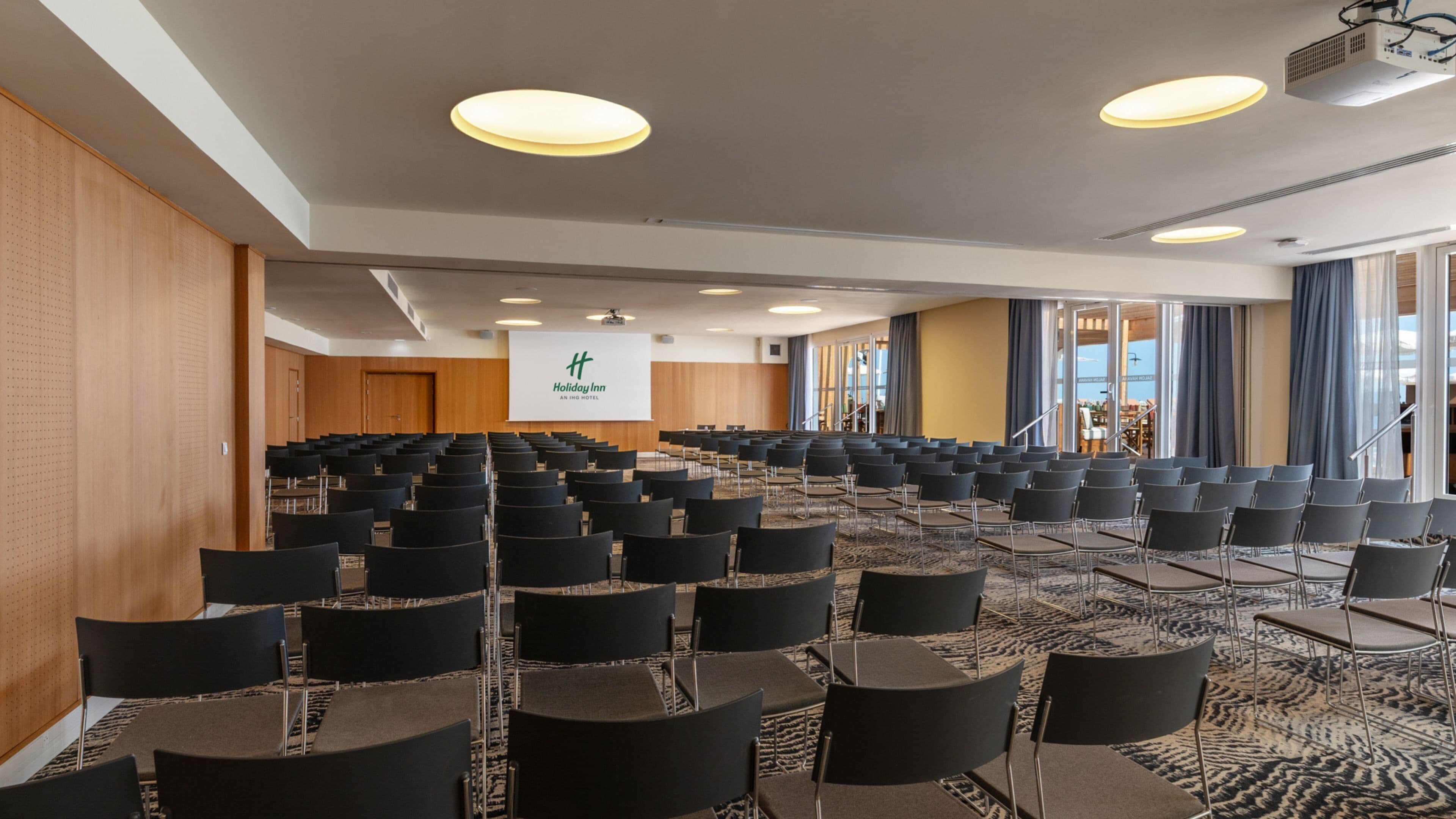 A conference room with rows of chairs, a projector screen, and a large window with curtains.