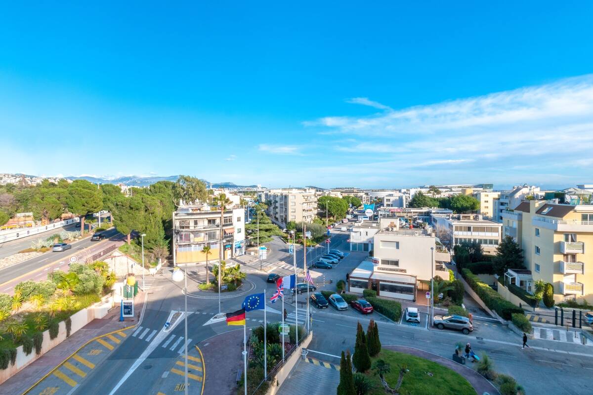 A cityscape with a mix of residential and commercial buildings, a clear blue sky, and a road with cars and pedestrians.
