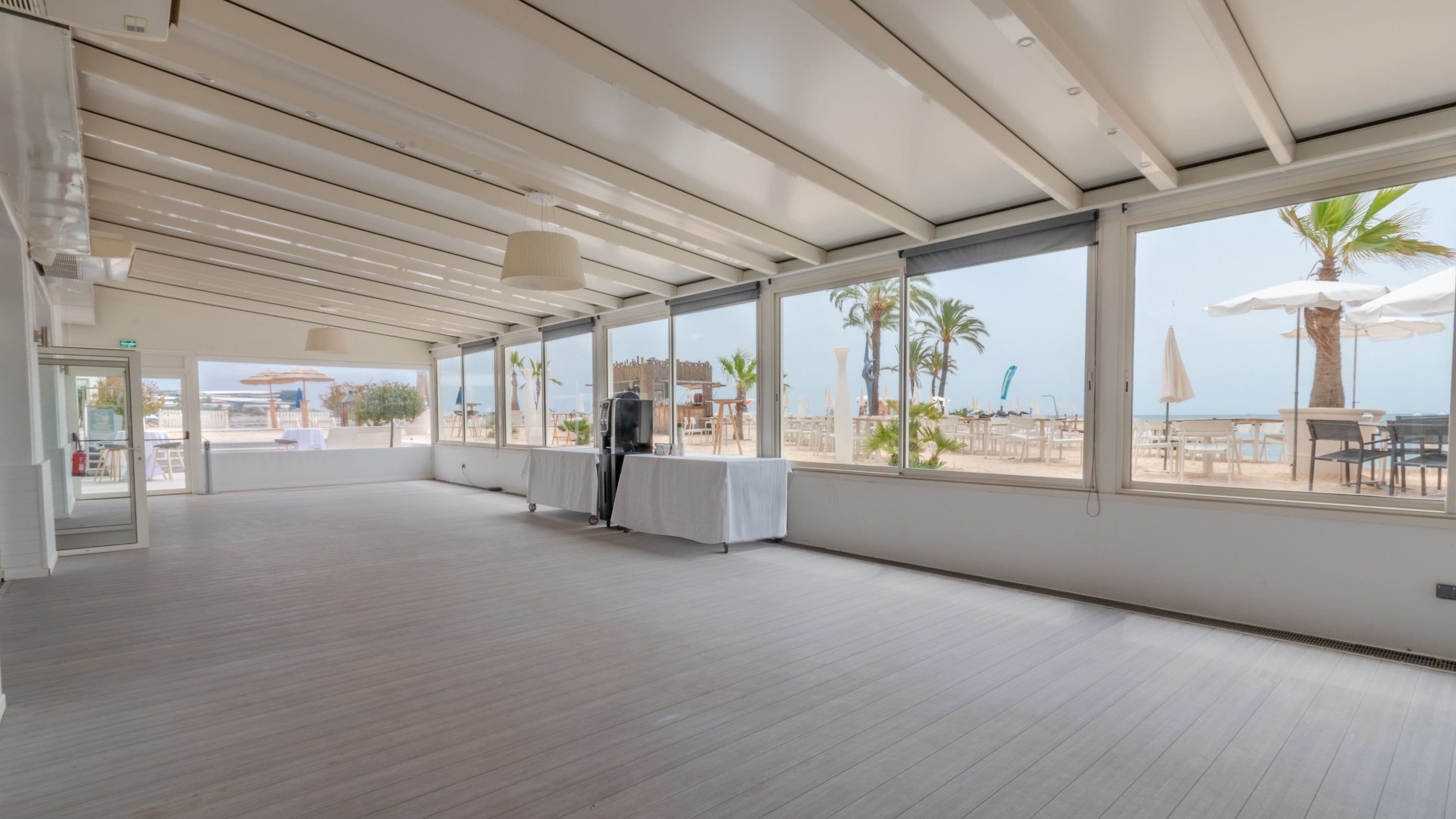 A spacious indoor area with large windows overlooking a beach, a grill, and a table set for dining.