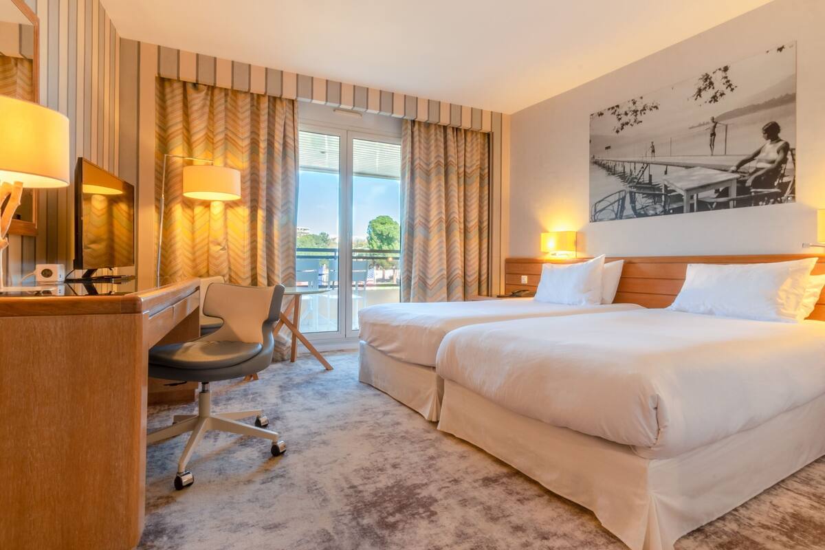 A hotel room with two beds, a desk, a chair, and a large window with curtains.