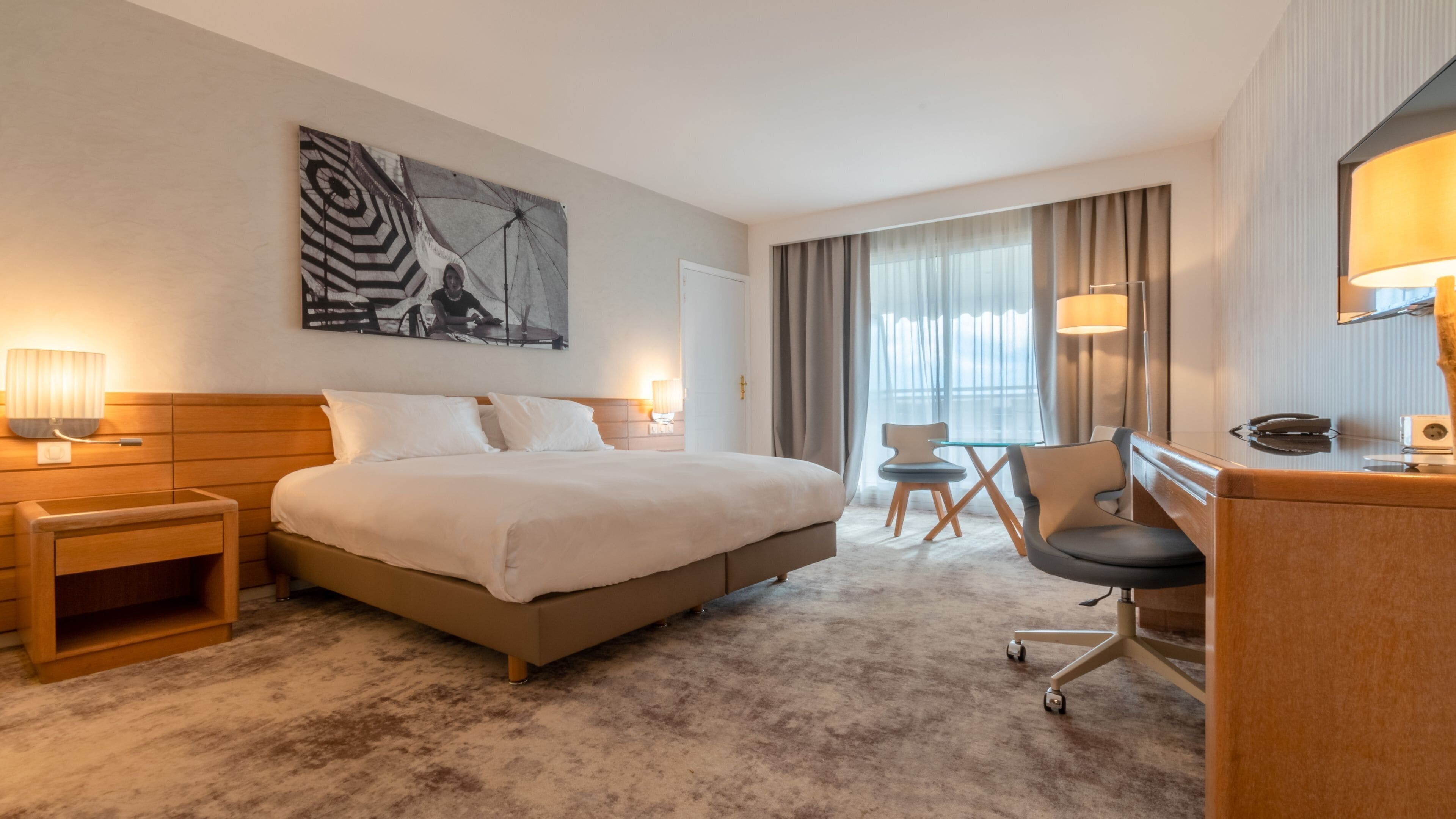A hotel room with a large bed, a desk, a chair, a small table, and a painting on the wall.