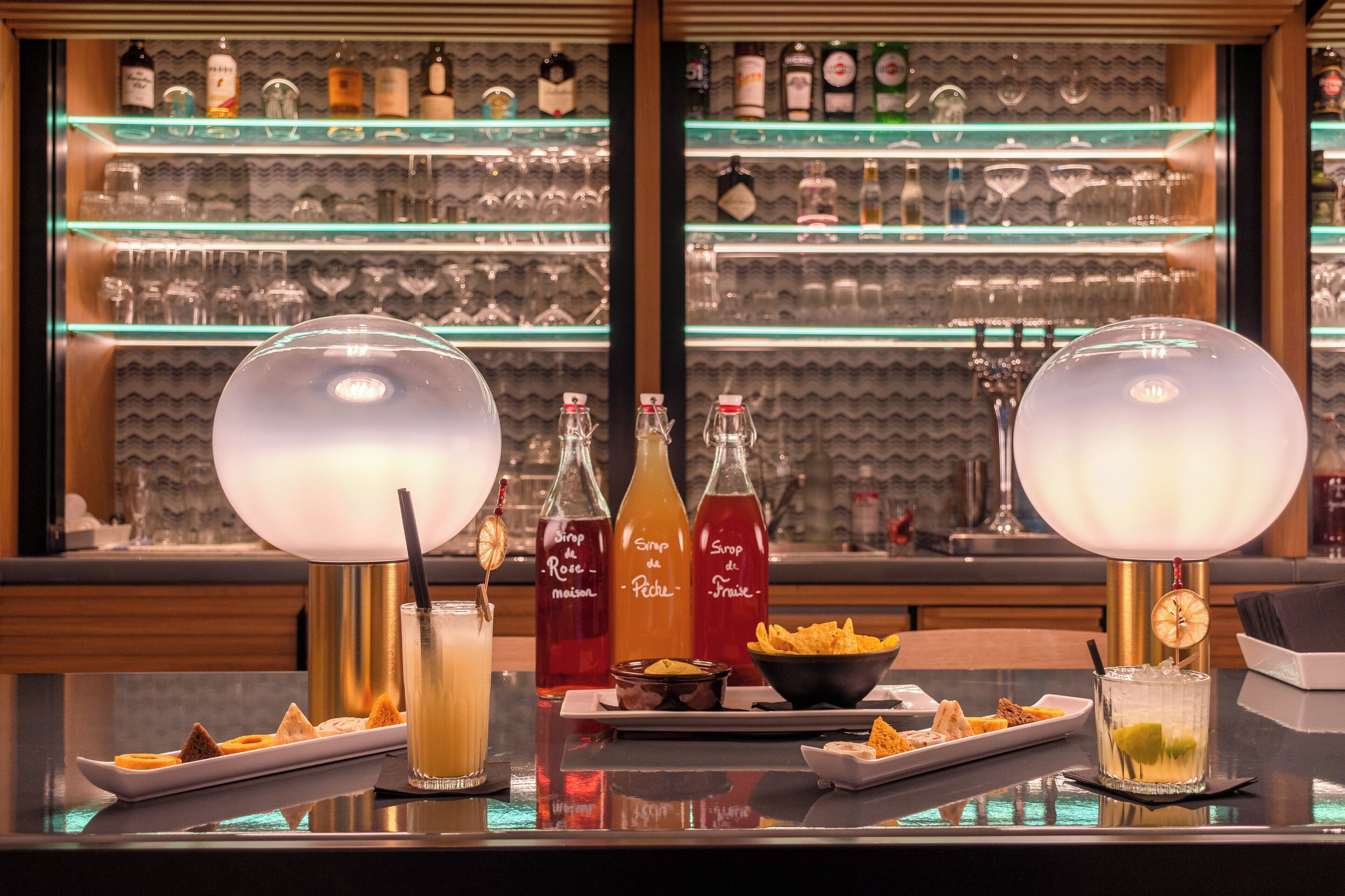 A bar area with two large lamps, a selection of drinks, and a display of bottles and glasses in the background.