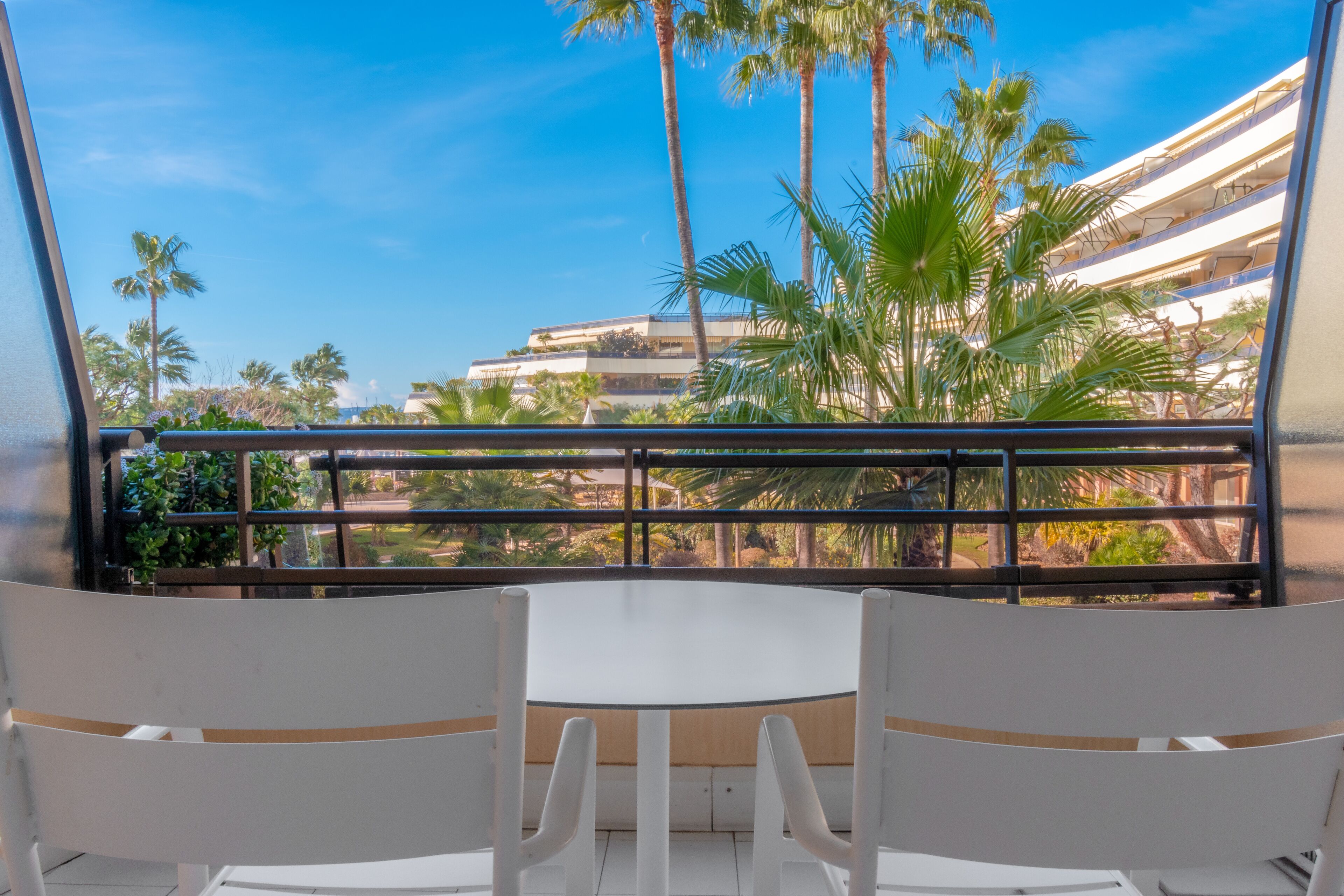 A balcony with a round table and two chairs, overlooking a palm tree-lined area with a building in the background.
