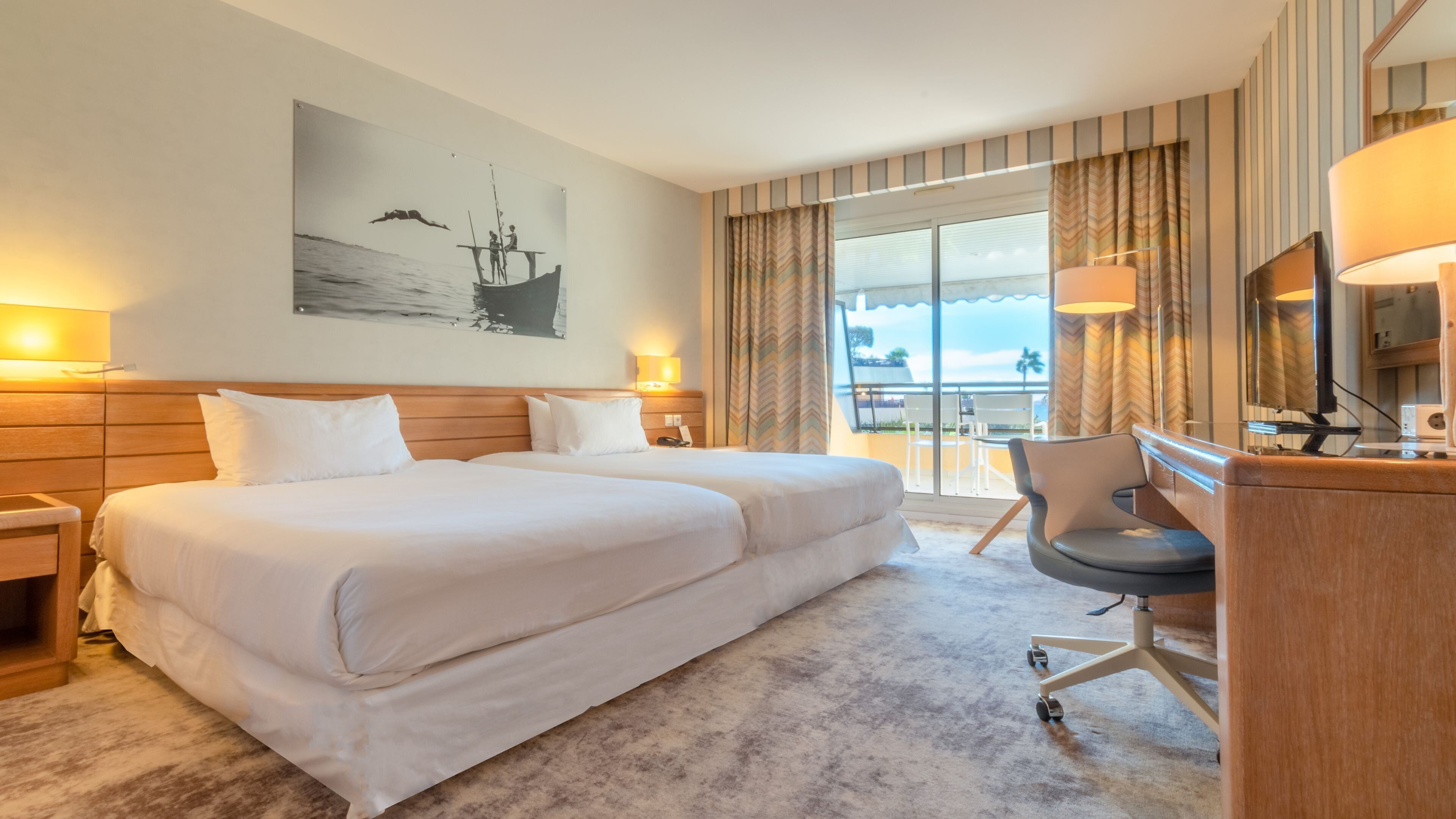 A hotel room with a large bed, a desk, and a chair. There is a window with curtains and a view of the outside.