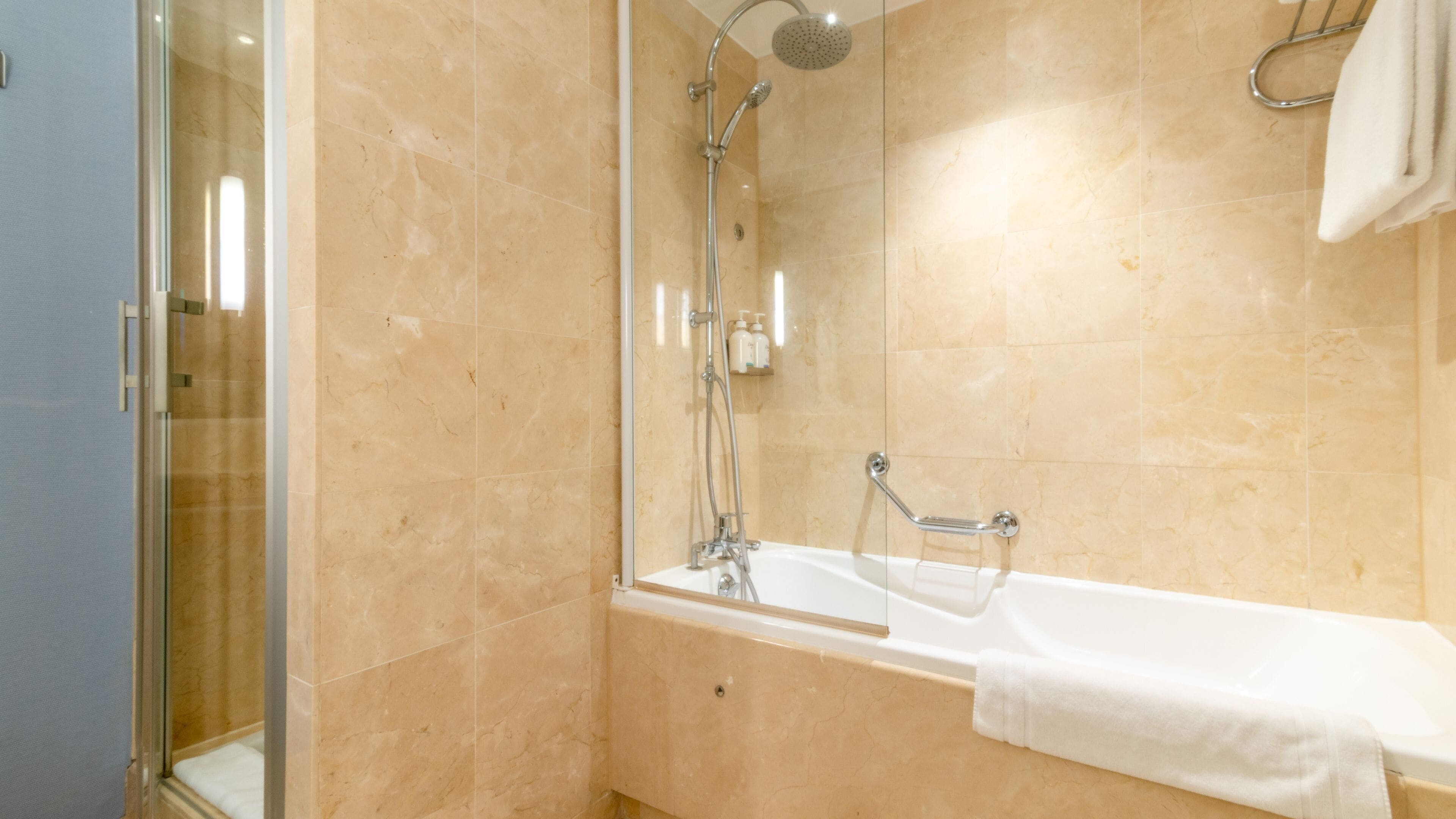 Une salle de bain moderne avec une douche à l’italienne en verre, une baignoire blanche et des carreaux de marbre beige.