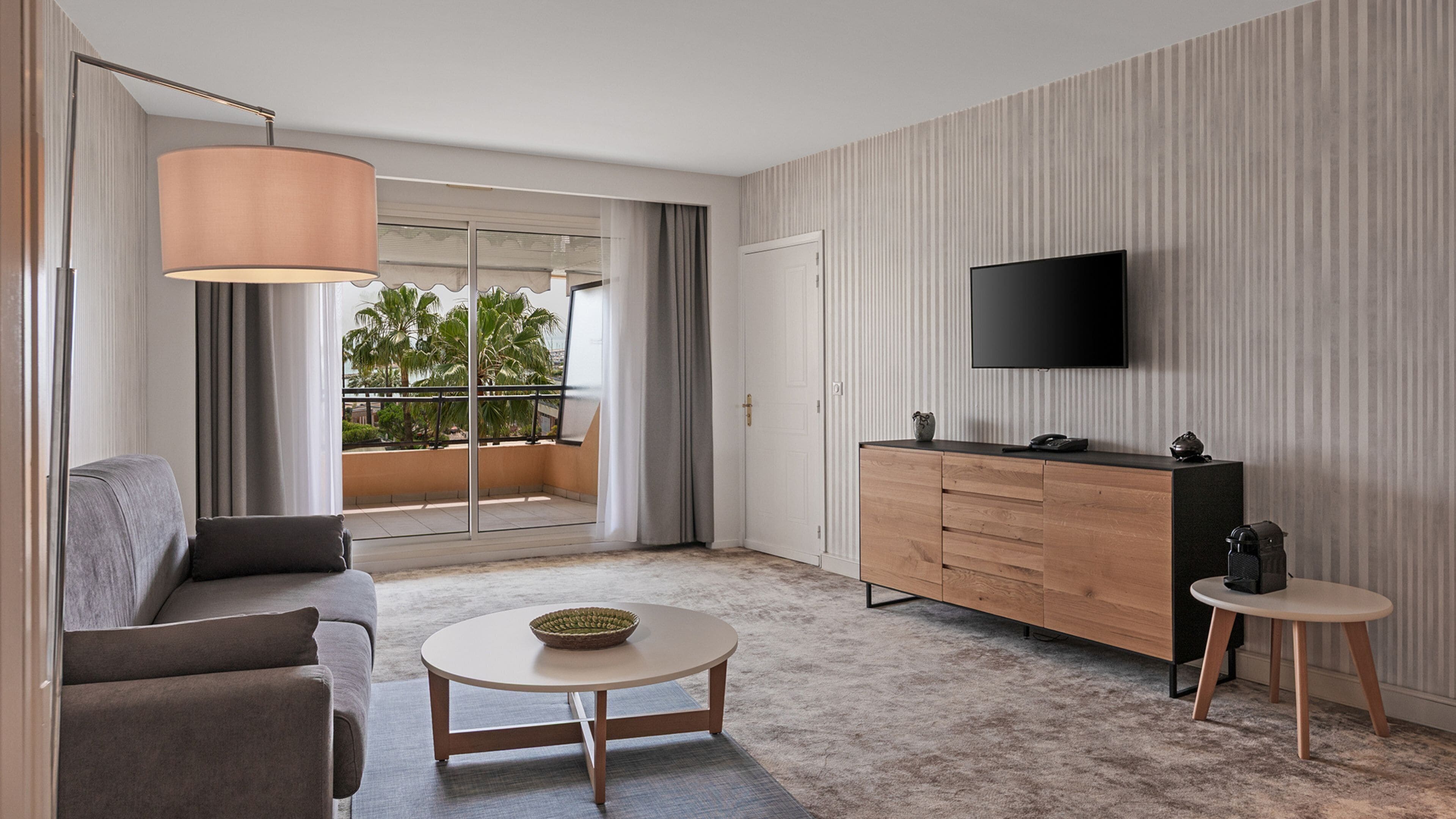 Une chambre d’hôtel moderne, dotée d’un canapé, d’une petite table ronde, d’une commode en bois, d’une télévision et d’un balcon offrant une vue sur les palmiers.