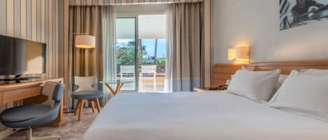A hotel room with a large bed, a desk with a television, a chair, and a balcony with curtains.