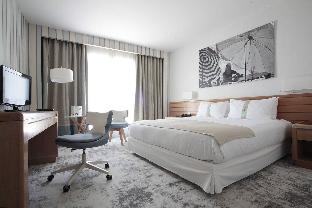 A hotel room with a bed, desk, chair, and a large window with curtains.