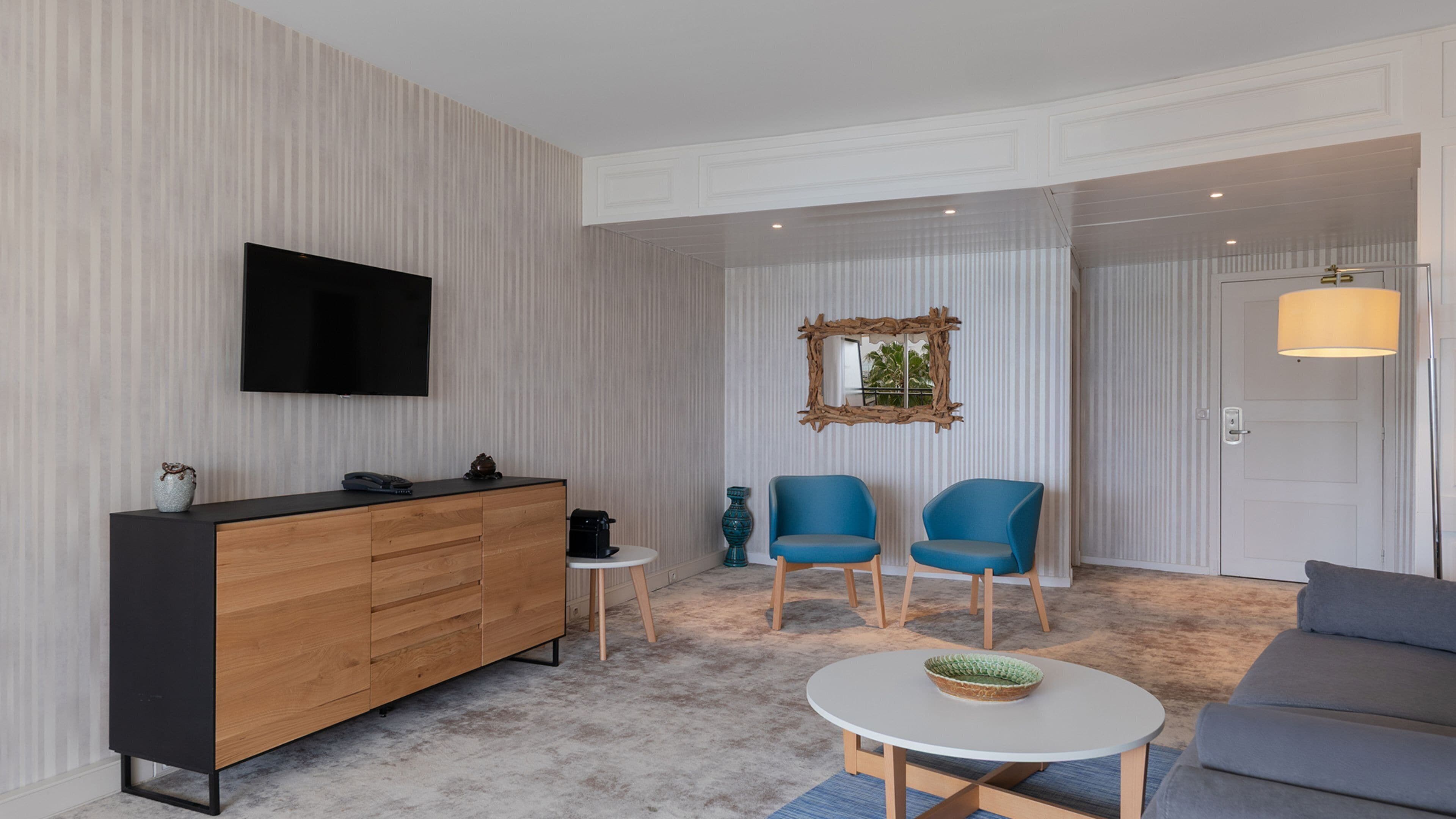 A modern living room with a wooden cabinet, two blue chairs, a grey sofa, a round coffee table, and a TV mounted on the wall.