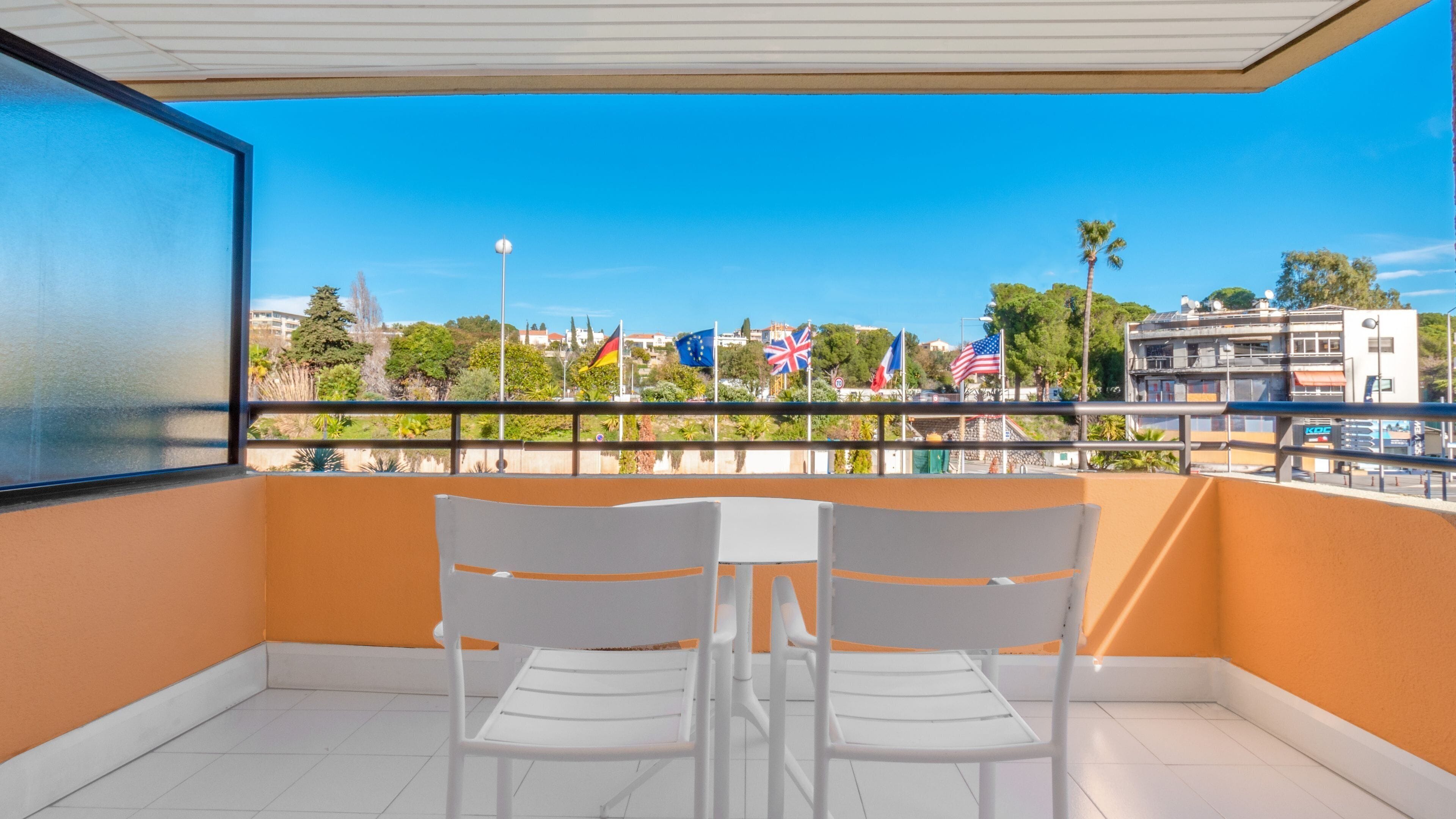 Un balcón con dos sillas blancas y una pequeña mesa, con vista a una calle con banderas y edificios.