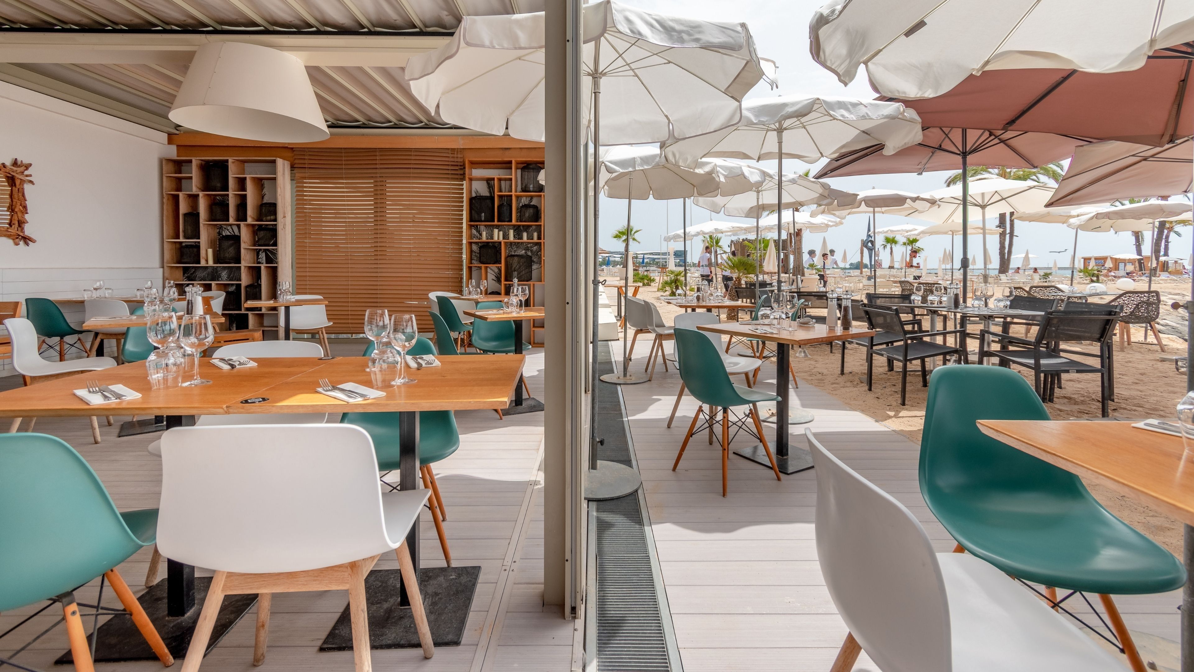 An outdoor dining area with wooden tables and chairs, umbrellas, and a view of the beach.