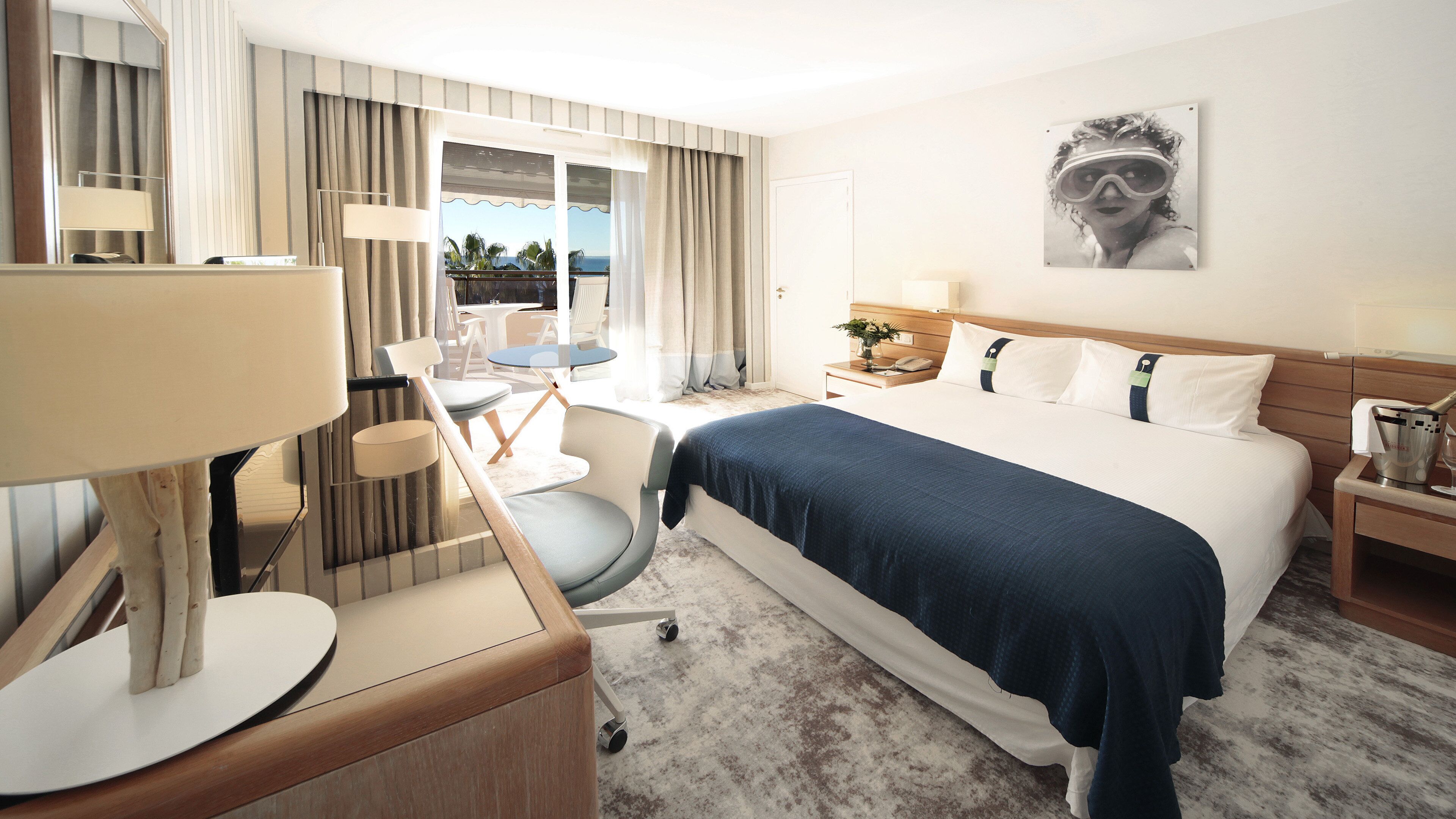 A hotel room with a bed, a desk with a lamp, a chair, a small table, and a window with curtains.