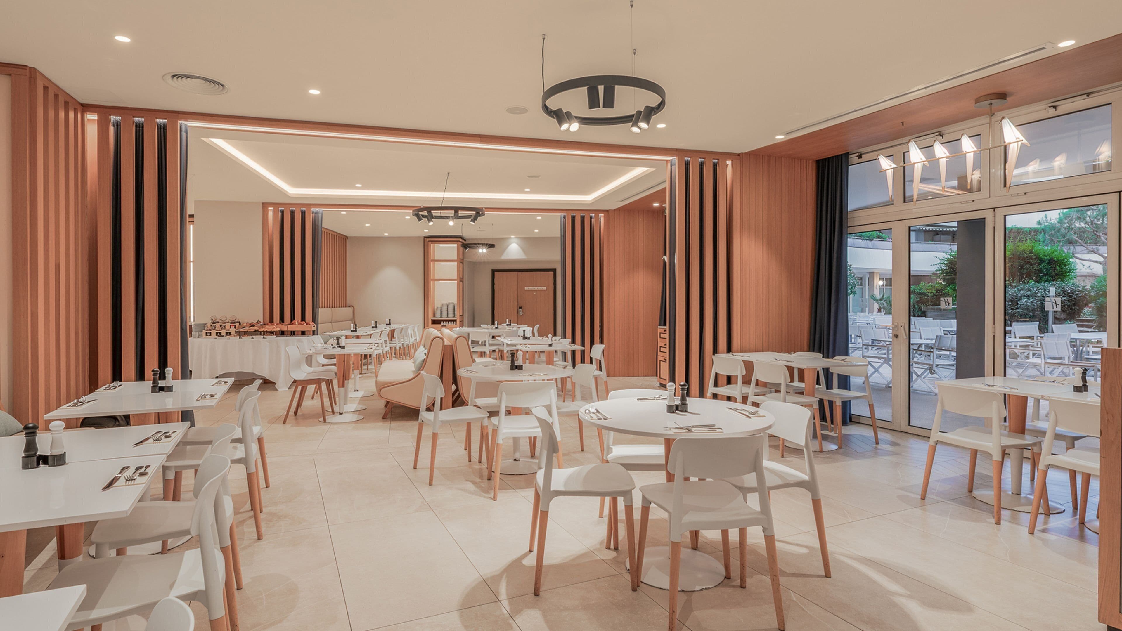 A modern dining area with multiple tables and chairs, large windows, and a view of the outdoors.