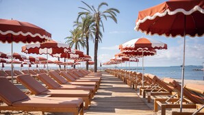 A beach with rows of wooden lounge chairs and red and white striped umbrellas. - Holiday Inn Nice-Port St Laurent by IHG (Saint-Laurent-du-Var)