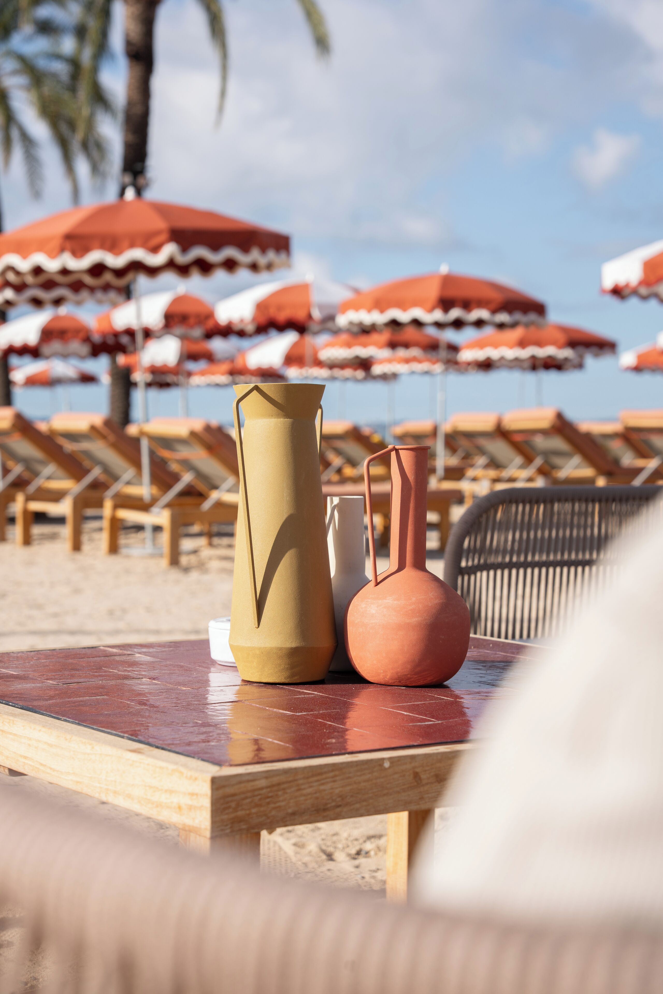 A beach setting with a table displaying two vases, beach chairs, and red umbrellas in the background.