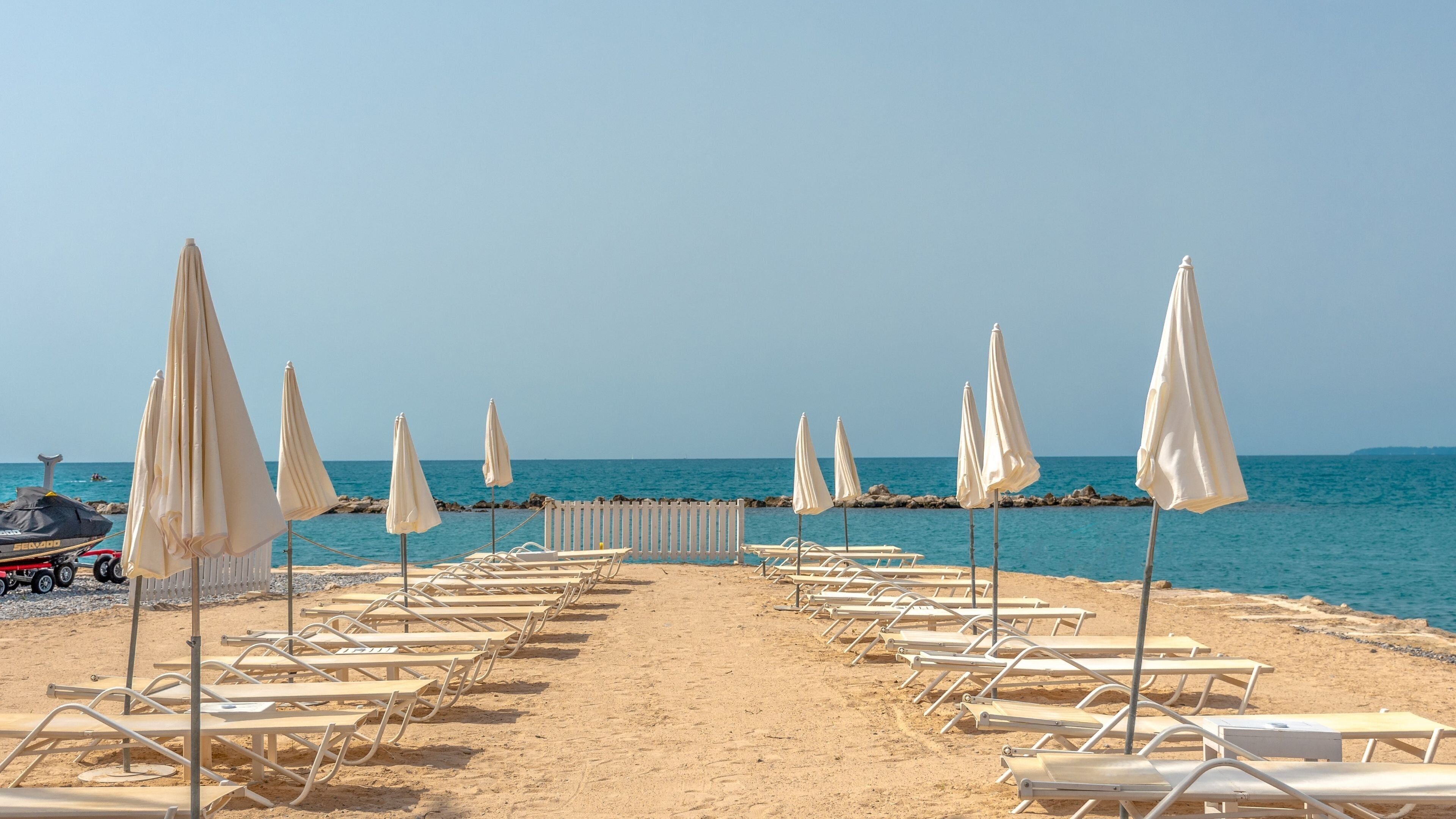 A beach with rows of sun loungers and umbrellas, a jet ski, and a rocky shoreline.