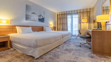 Habitación de hotel con una cama grande, dos mesitas de noche con lámparas, un escritorio con silla y un amplio ventanal con cortinas.