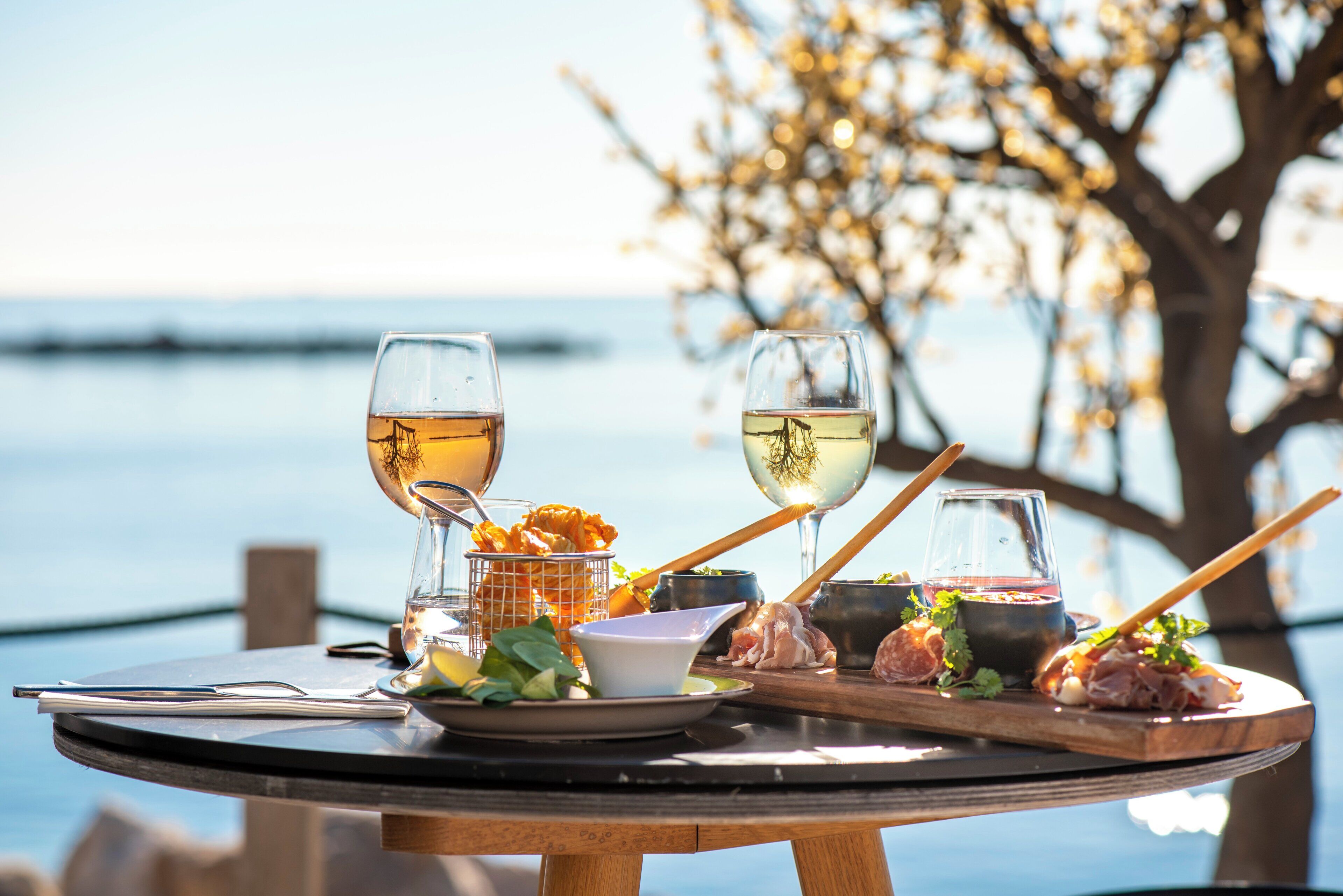 Een ronde tafel met uitzicht op de zee, een glas witte wijn en diverse soorten eten.