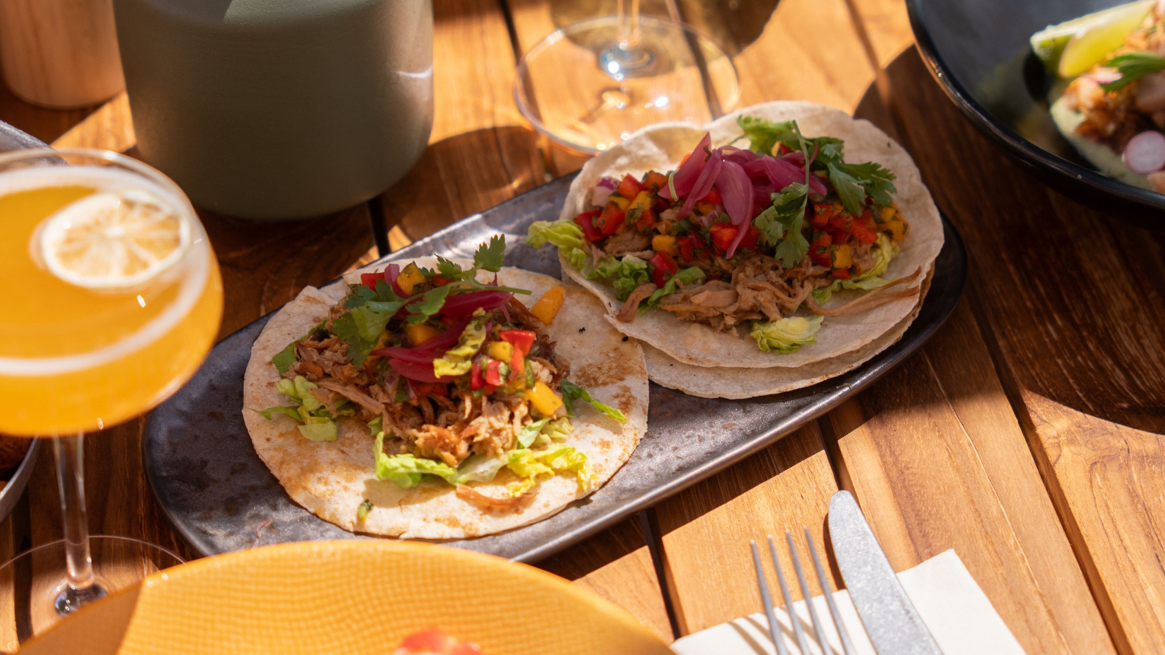 Two tacos on a metal tray with a glass of orange juice and a glass of wine on a wooden table.