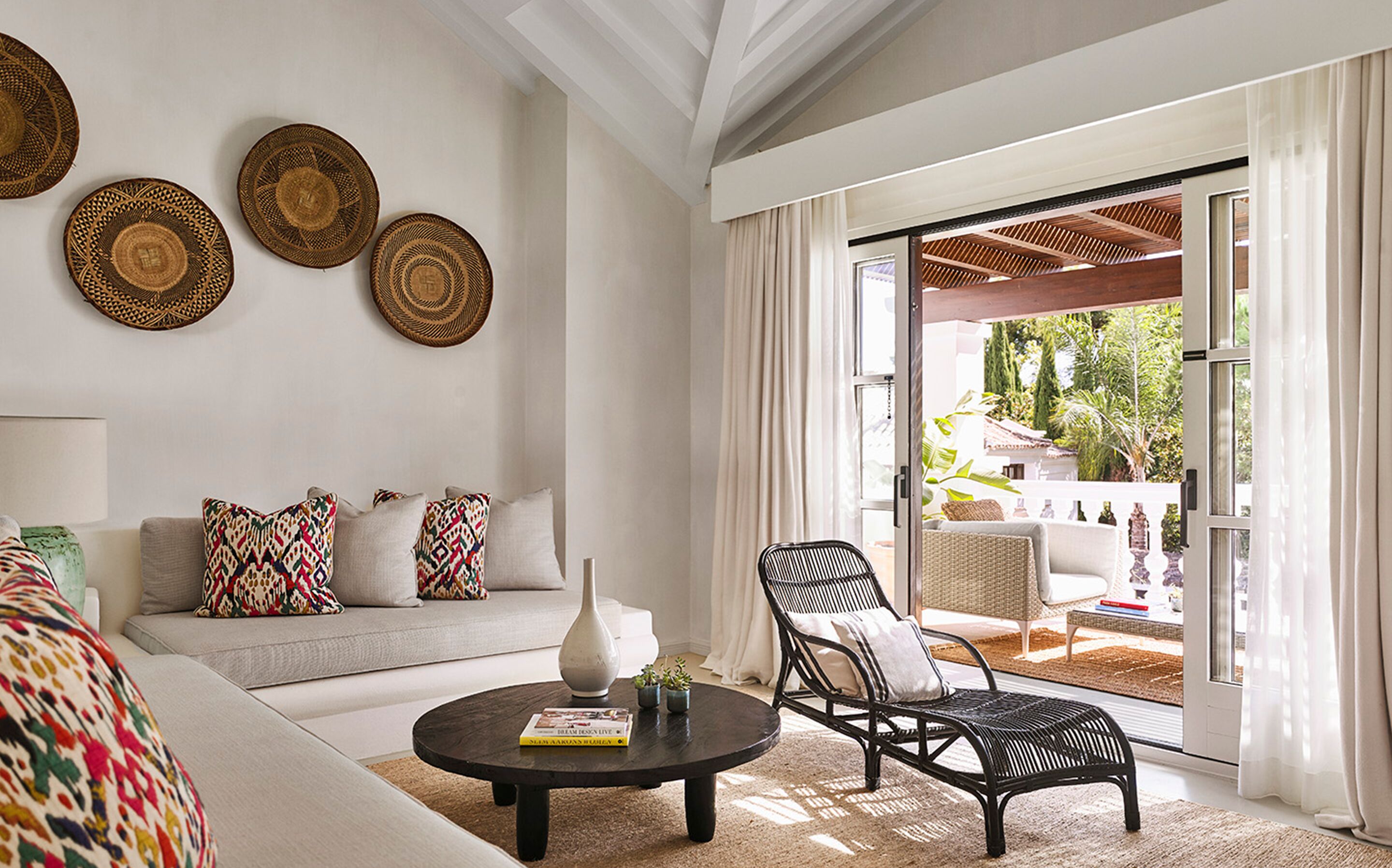 A living room with a sofa, coffee table, and a chair. There are woven baskets on the wall and a view of the outdoors through a sliding glass door.