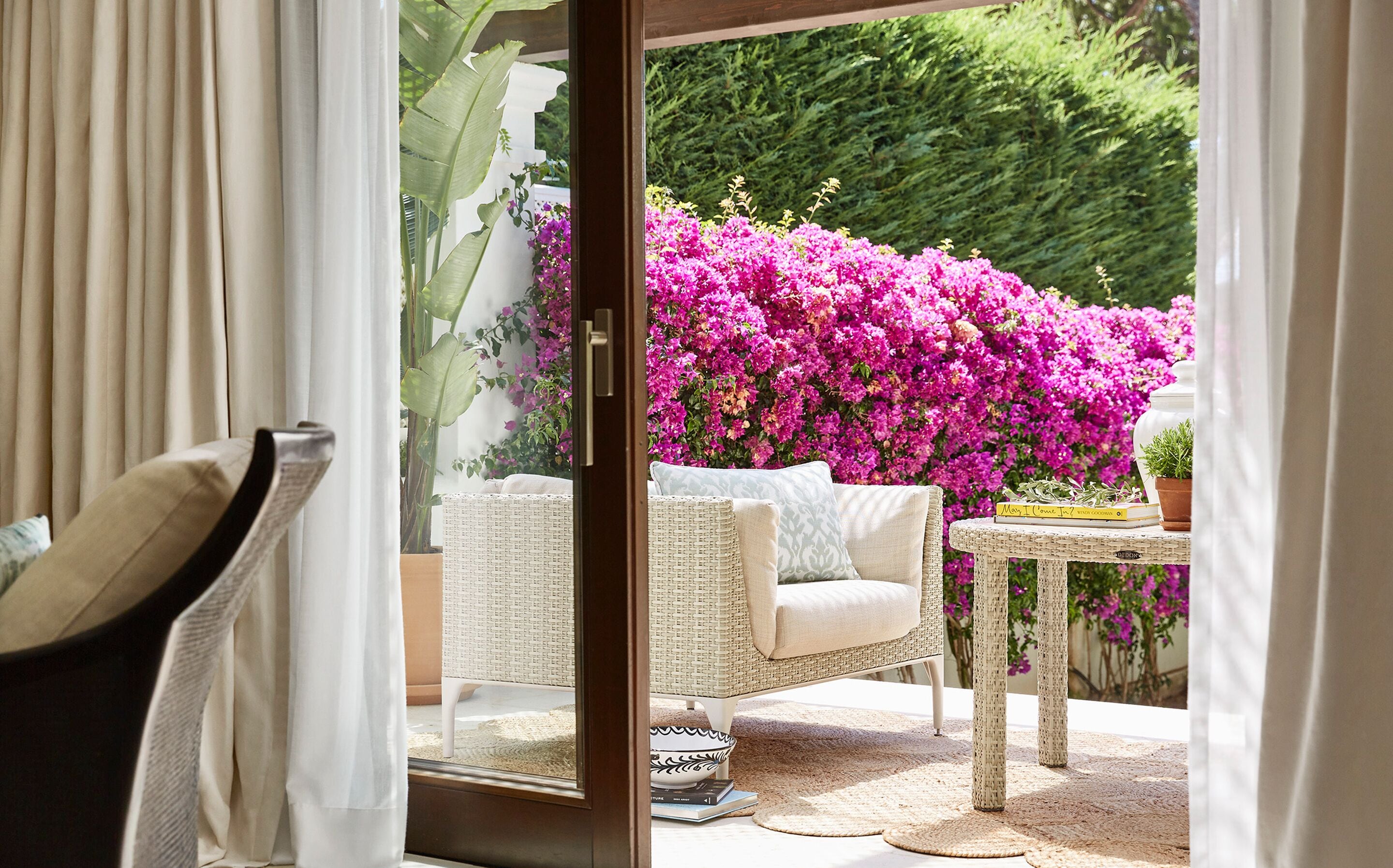 A wicker chair and a small table with a book and a potted plant in a sunny outdoor area with a view of vibrant pink flowers and greenery.