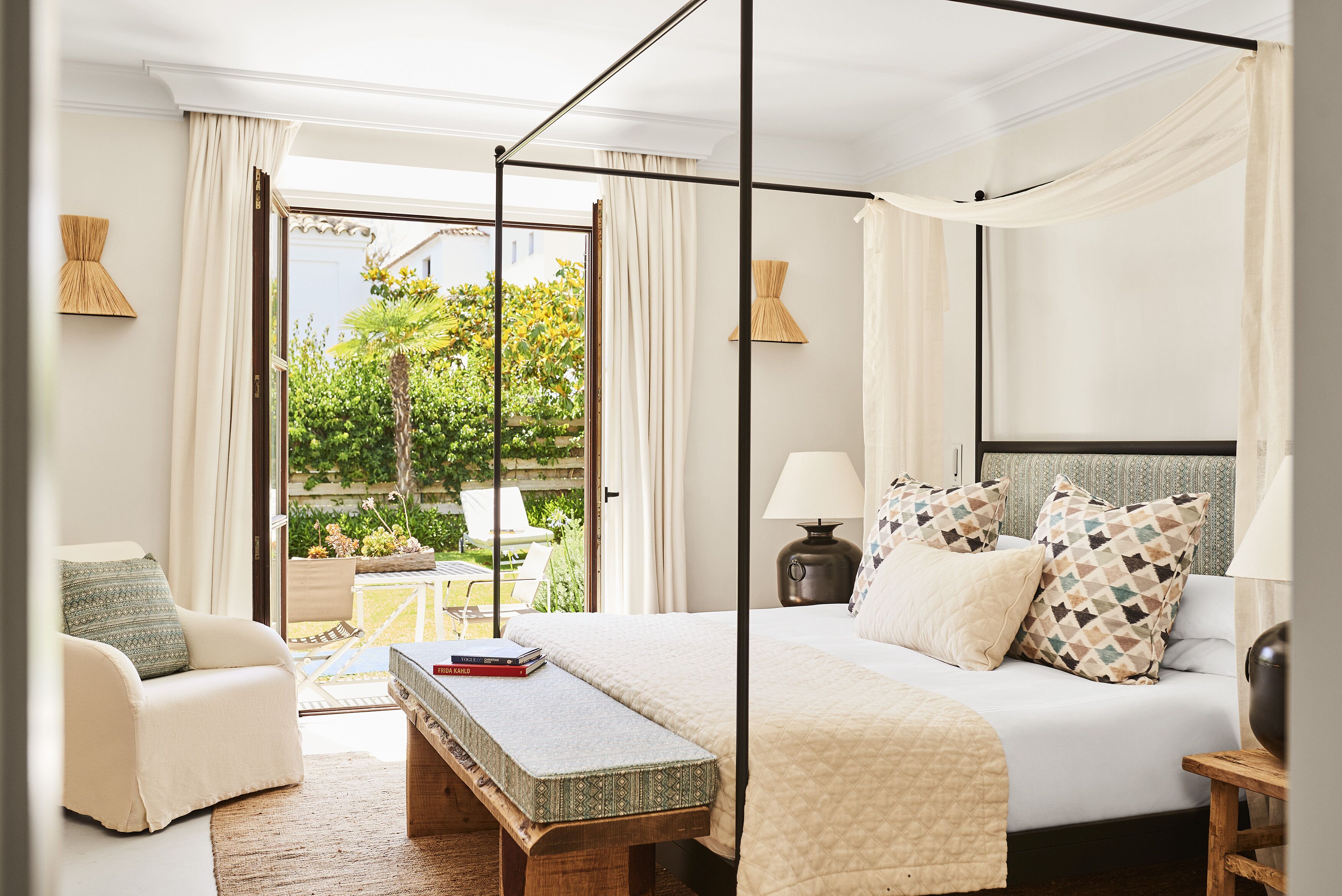 A bedroom with a four-poster bed, a bedside table, a chair, and a view of the garden through a glass door.