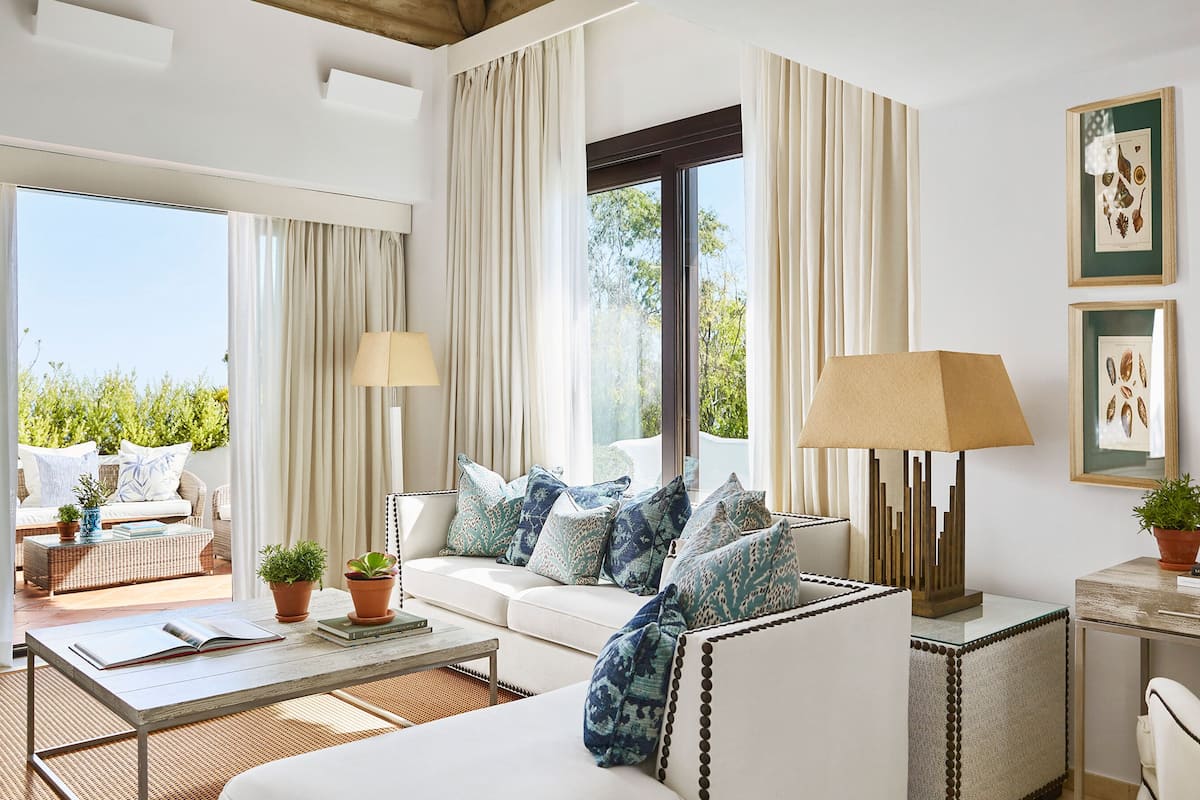 A modern living room with a sofa, a coffee table, and a lamp. There are potted plants and a framed picture on the wall.