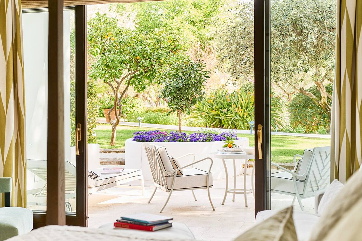 A room with a sliding glass door leading to a garden with a table, chairs, and a small tree.