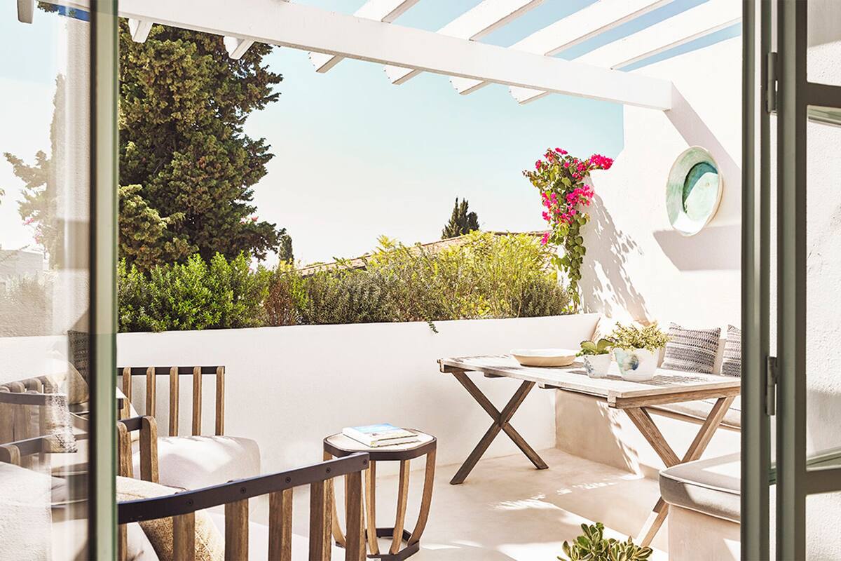 A sunlit patio with a wooden table, chairs, and a small round table with books and a plant.