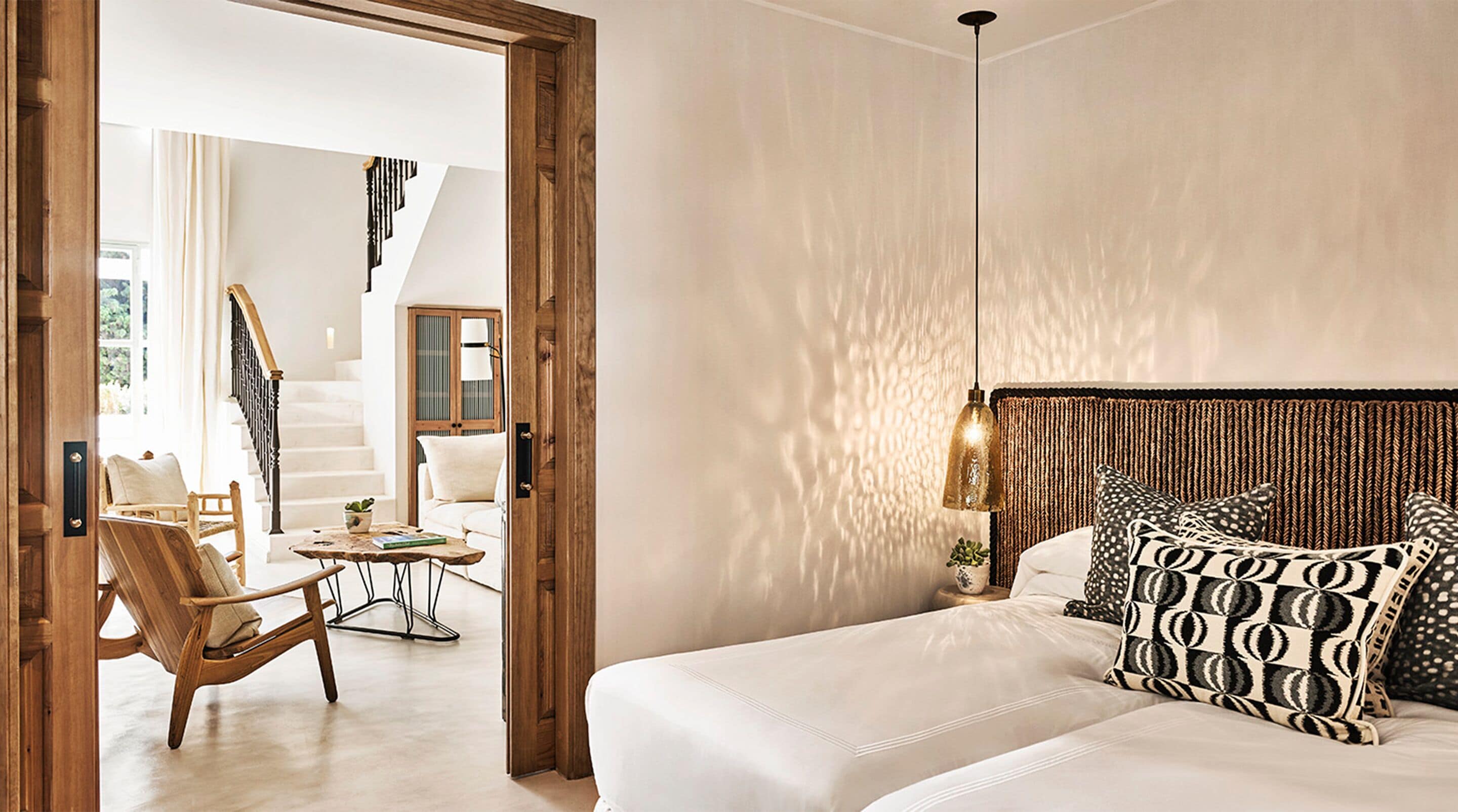 A bedroom with a bed, patterned pillows, a hanging light fixture, and a wooden door leading to a living area with a chair and a table.