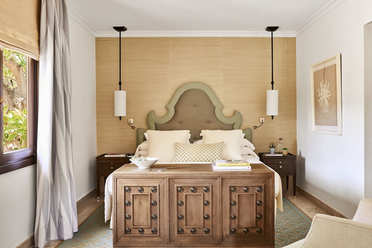 A bedroom with a wooden headboard, two bedside tables, a bed with pillows, a nightstand with a lamp, and a window with curtains.